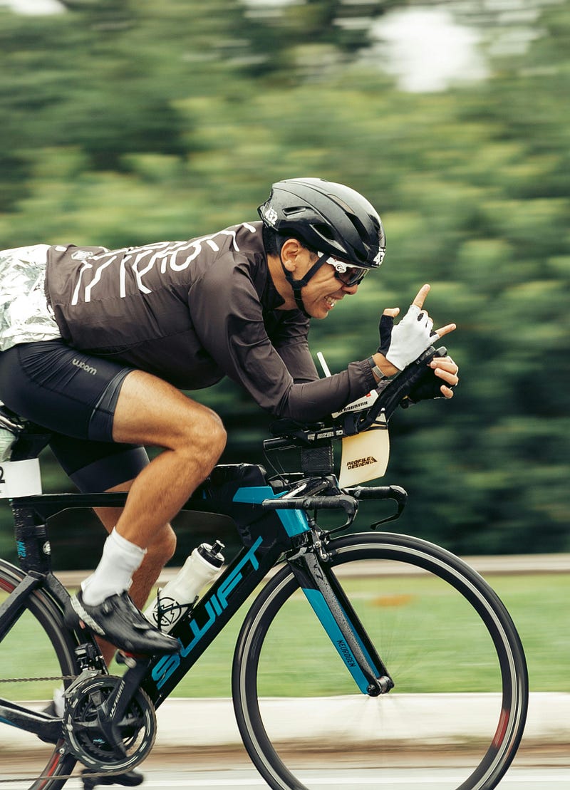 A happy man riding a racing cycling bike whilst passing by the camera at a fast speed holding up the rock sign with his right hand.